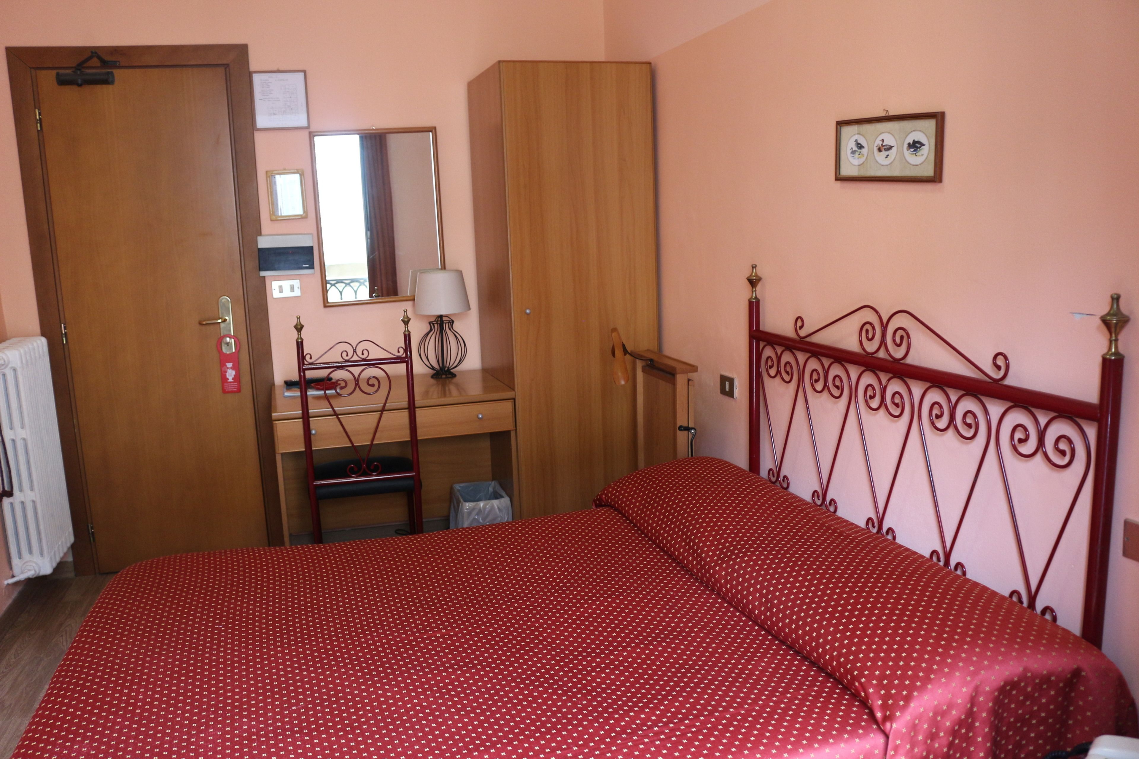 A bedroom with a bed, a wooden wardrobe, a desk with a chair, a mirror, and a radiator.