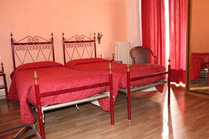 Una cama doble con estructura metálica y ropa de cama roja, una silla de madera y una cortina roja.
