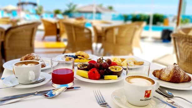 Une table dressée pour le petit-déjeuner, avec des croissants, des fruits et du café, et une vue sur la plage.