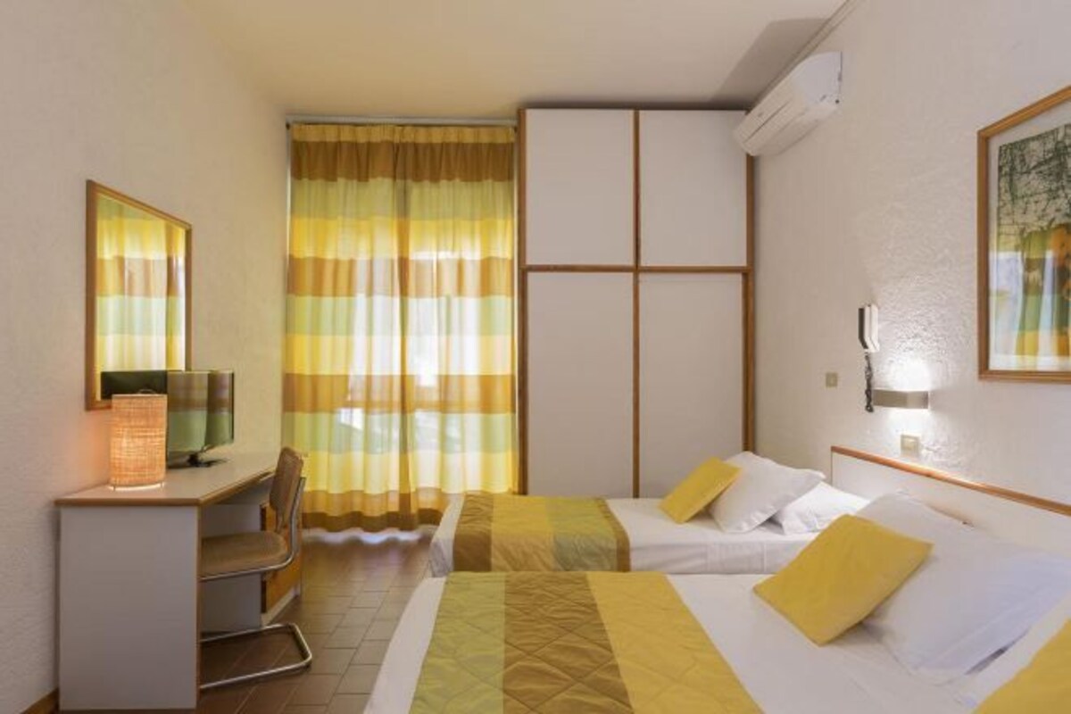 A hotel room with a desk, two beds, a chair, a television, and a window with curtains.