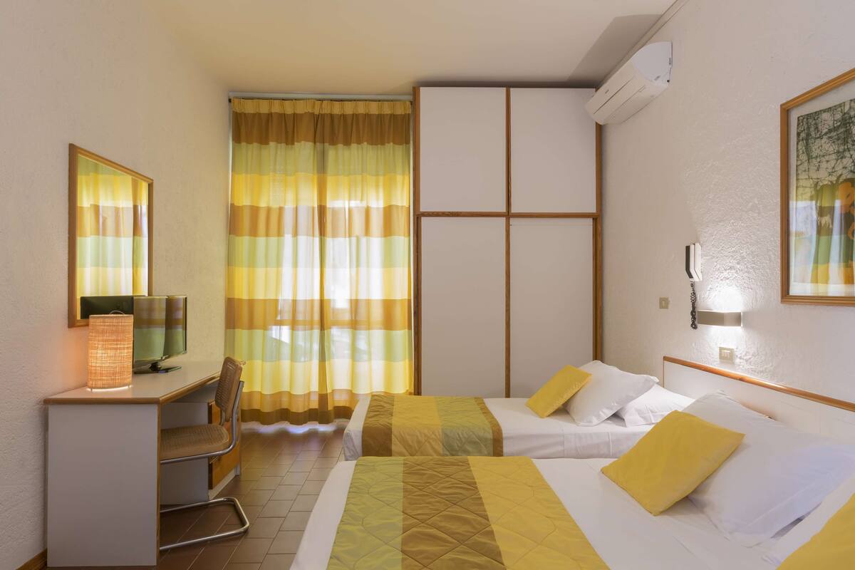 A hotel room with two beds, a desk with a computer, a chair, and a large window with curtains.