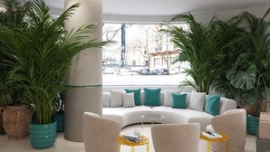 Un salon moderne avec un canapé incurvé, deux fauteuils et une table basse. On y trouve de grandes plantes en pot et un pilier imposant.