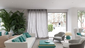 Un salon moderne avec un canapé blanc, des coussins turquoise, un fauteuil gris et une table basse sur laquelle sont posés des livres. On y trouve également des plantes en pot et, grâce aux grandes fenêtres, une vue sur la rue.