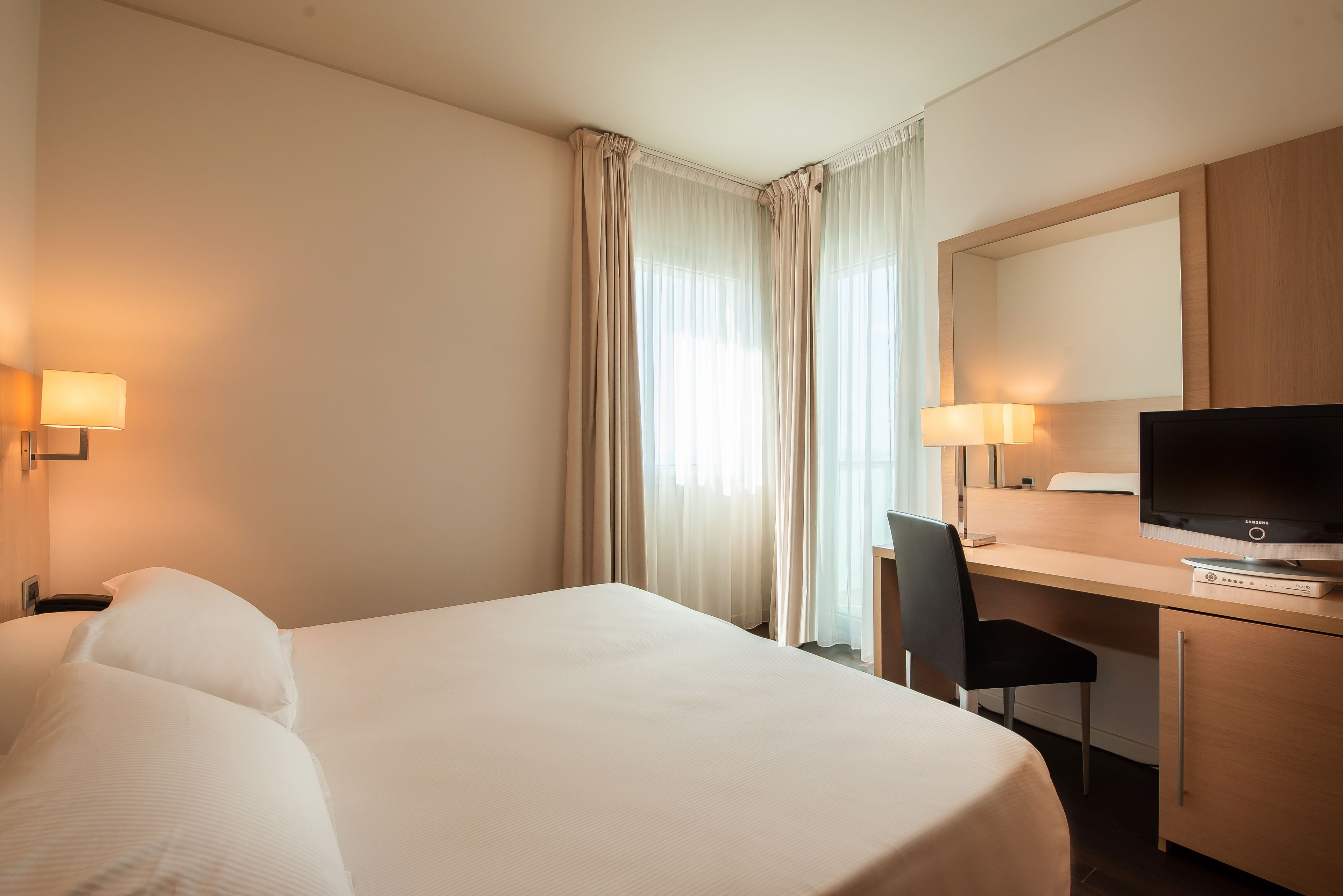 A hotel room with a bed, a desk with a TV, a chair, and two lamps.