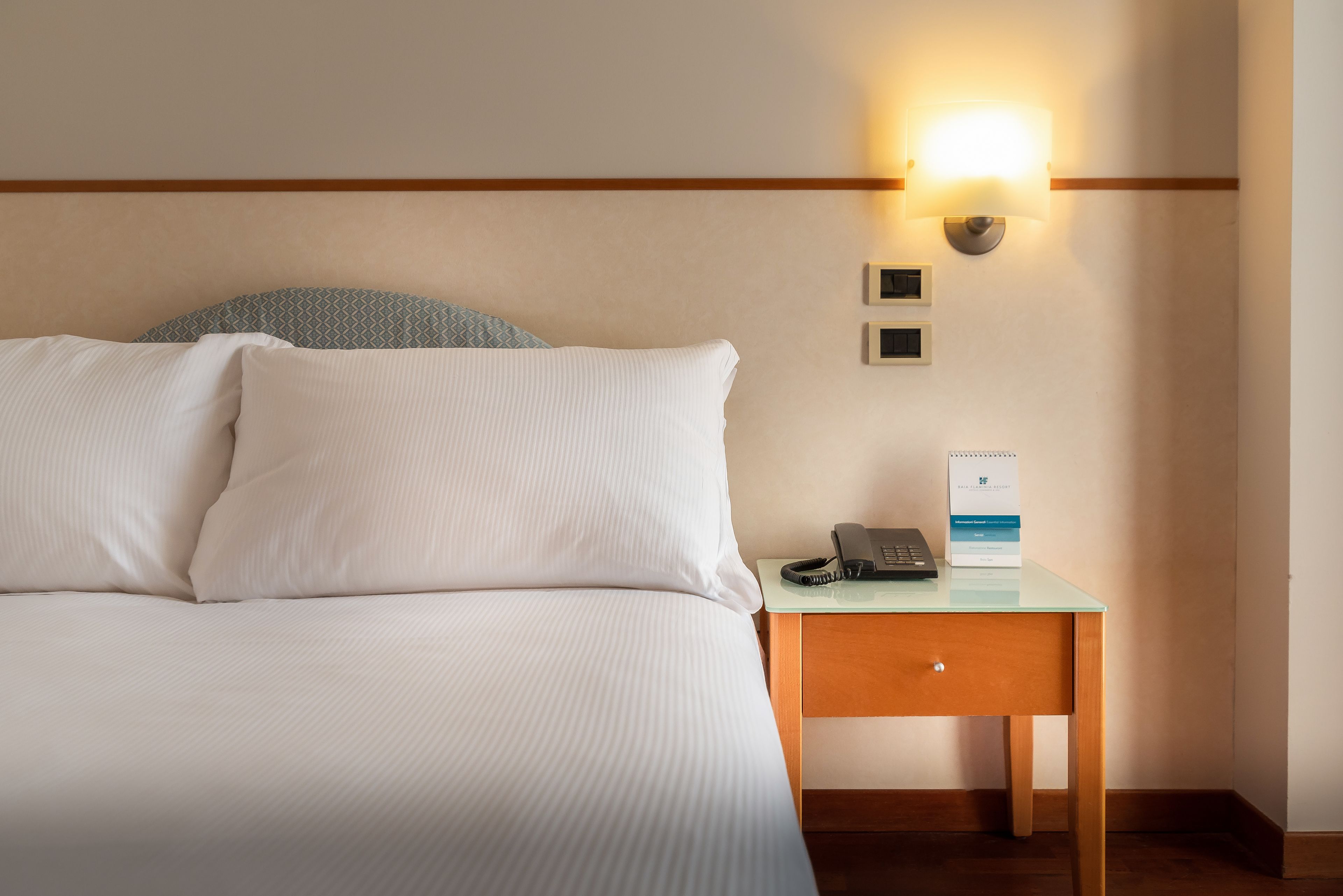 Ein Hotelzimmer mit einem Bett, einem Nachttisch mit Telefon und Schild sowie einer Wandleuchte.