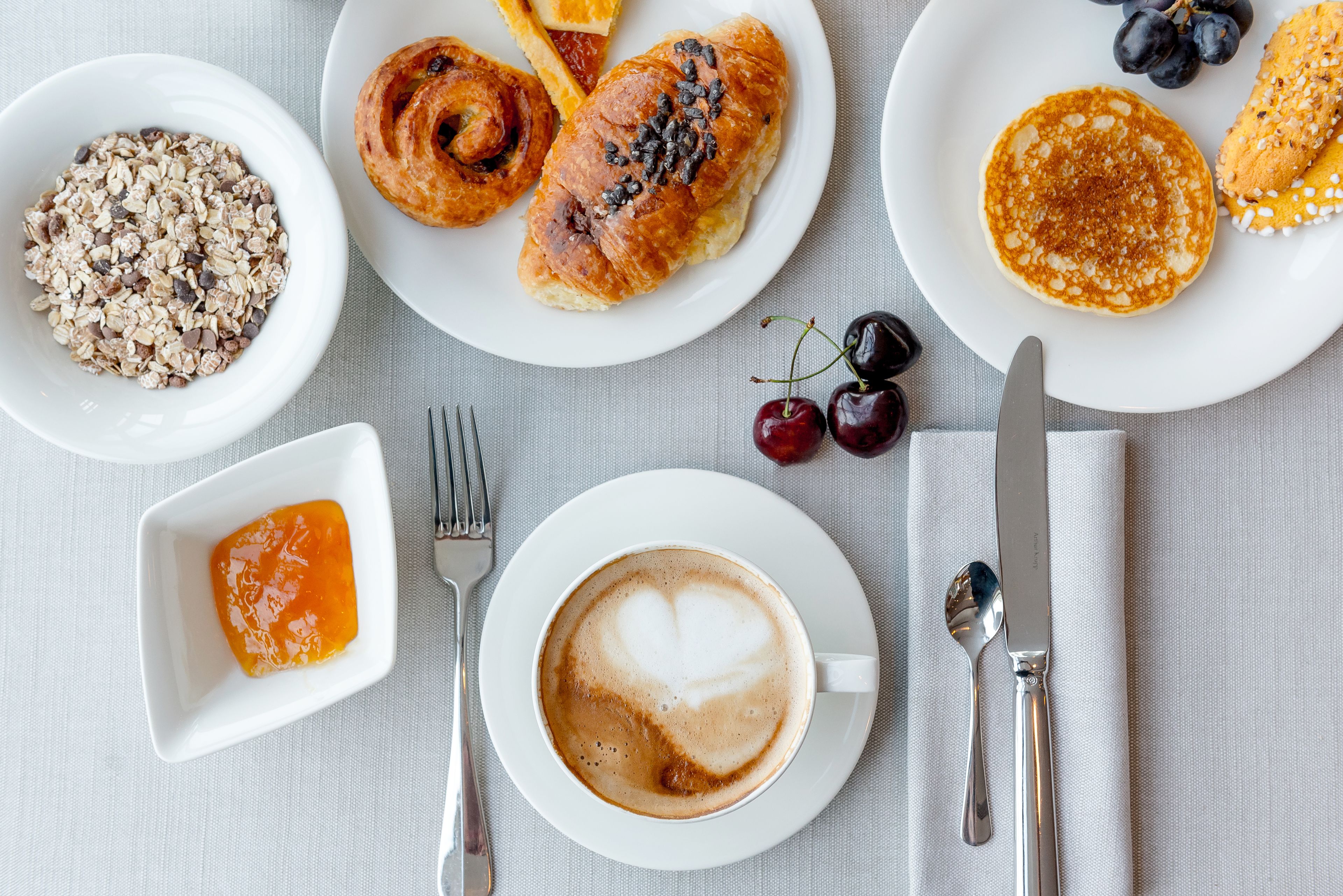 En frukostuppdukning med croissanter, pannkakor och en kopp kaffe med vackert hjärtformat mönster i mjölkskummet.