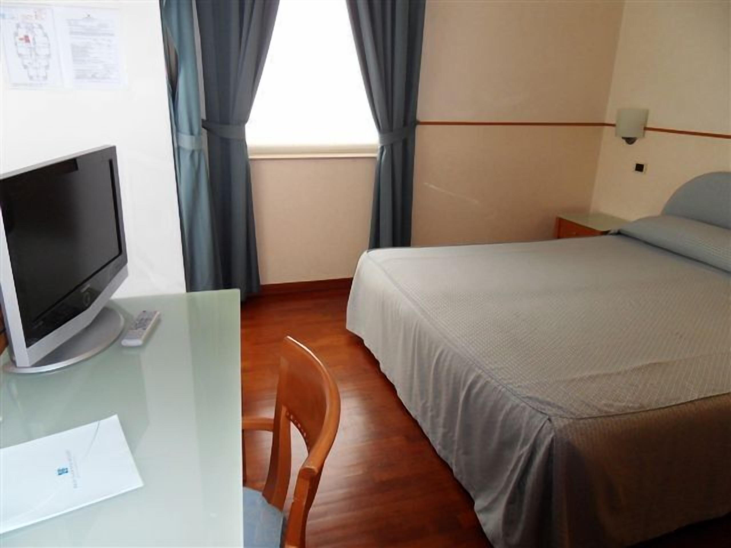Une chambre d’hôtel avec un lit, un bureau avec un ordinateur, une chaise et une fenêtre avec des rideaux.