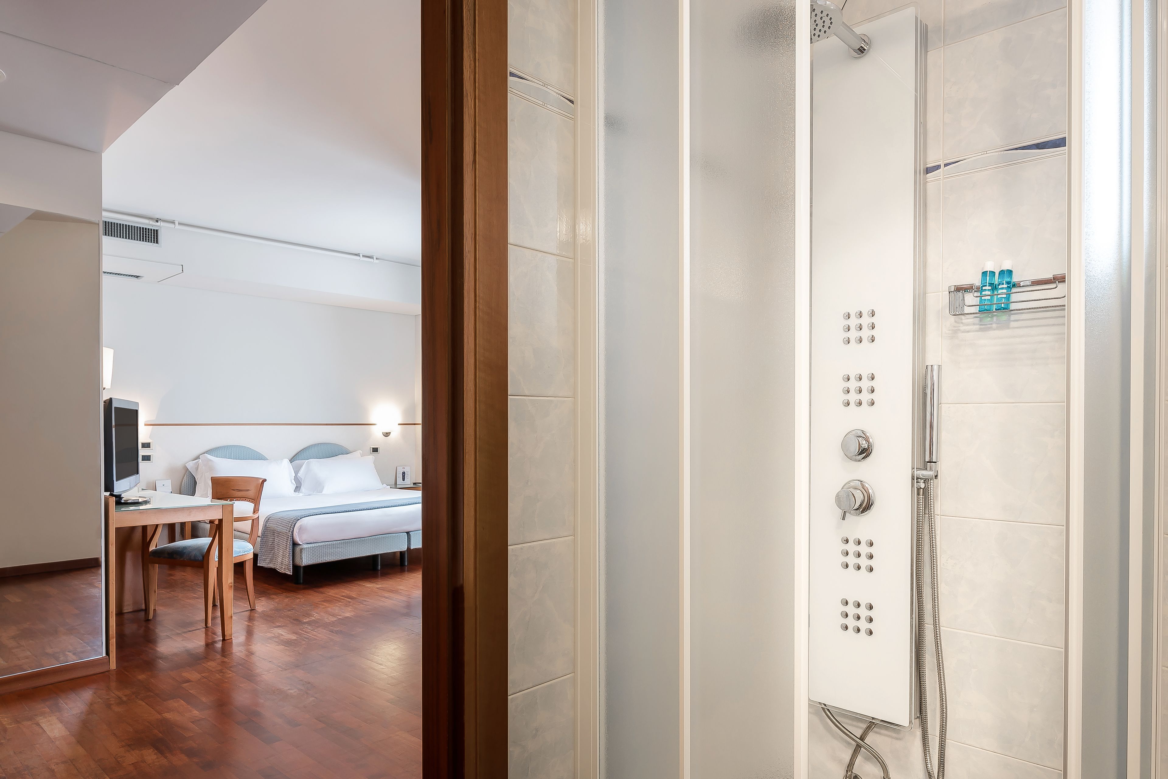 A hotel room with a wooden desk, a bed with white linens, and a shower area with a control panel.