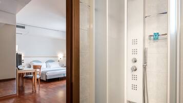A hotel room with a wooden desk, a bed with white linens, and a shower area with a control panel.
