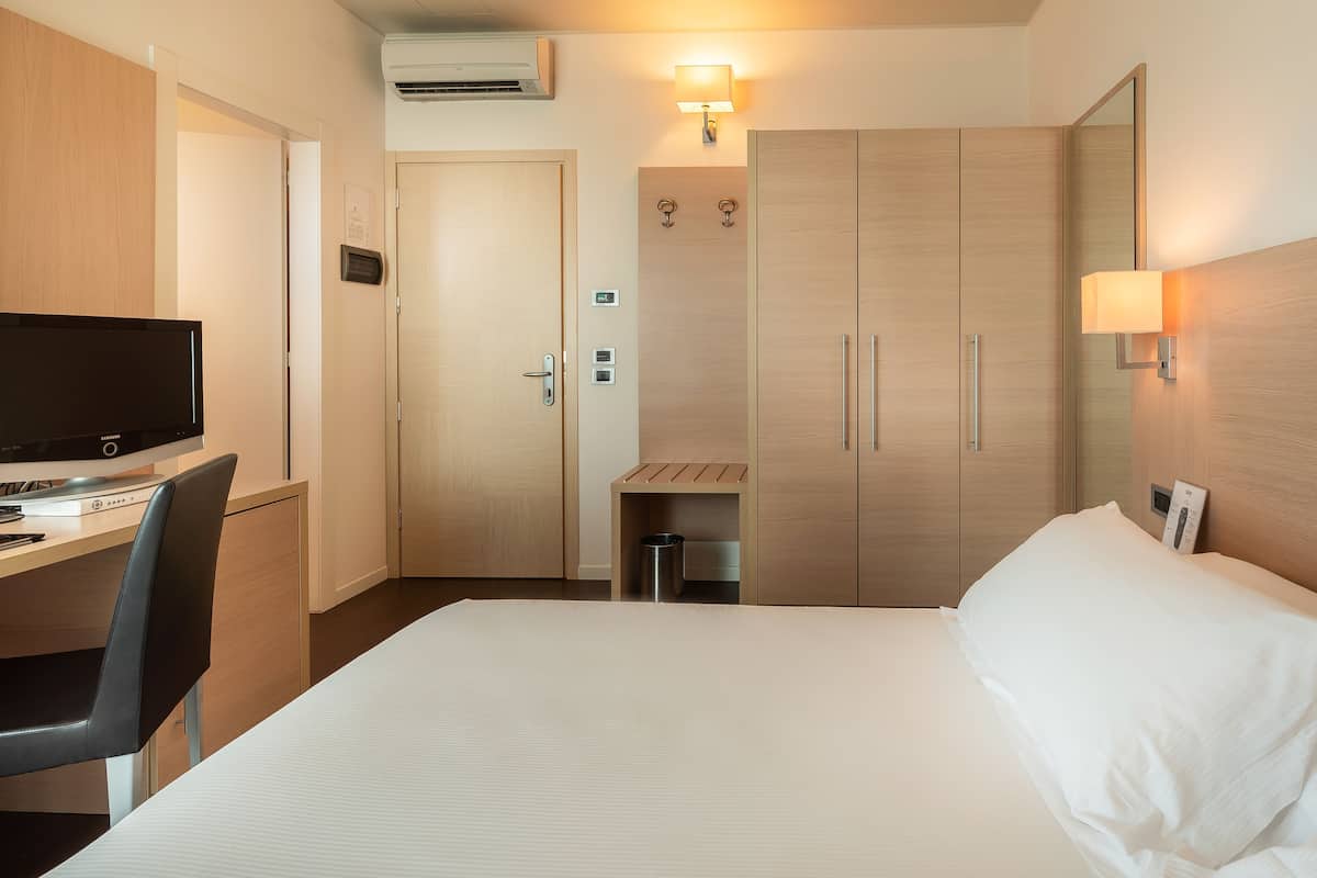 Habitación de hotel con cama, escritorio con computadora, televisor, armario y un pequeño bote de basura.