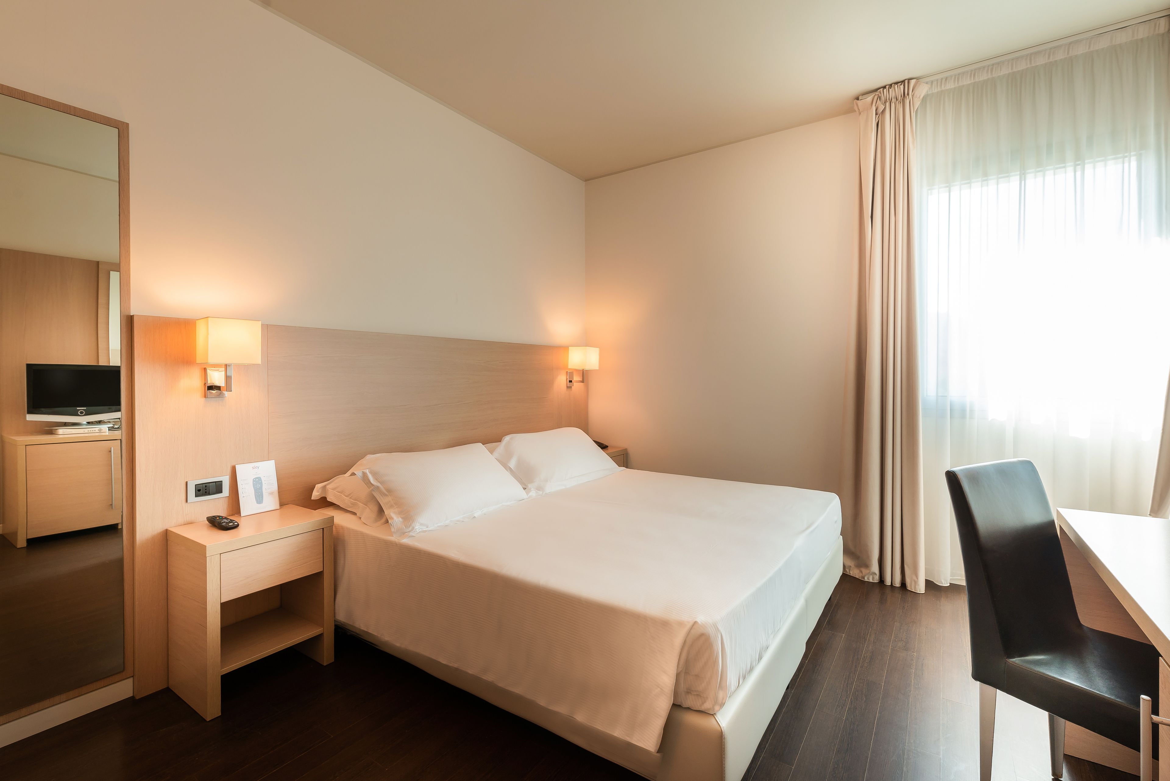 Ein Hotelzimmer mit einem großen Bett, Nachttischlampen, einem Schreibtisch, einem Sessel, einem Fernseher und einem Spiegel.