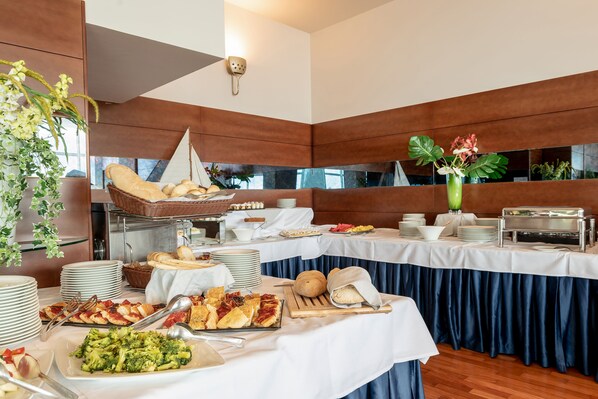 Montaje de bufé con diversos alimentos, entre ellos pan, pastelería y verduras, dispuestos sobre mesas con manteles blancos y cortinas azules.