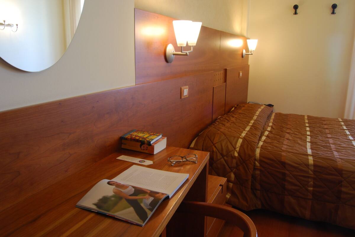 A hotel room with a wooden headboard, a bed with a brown quilt, a desk with a book, glasses, and a notebook.