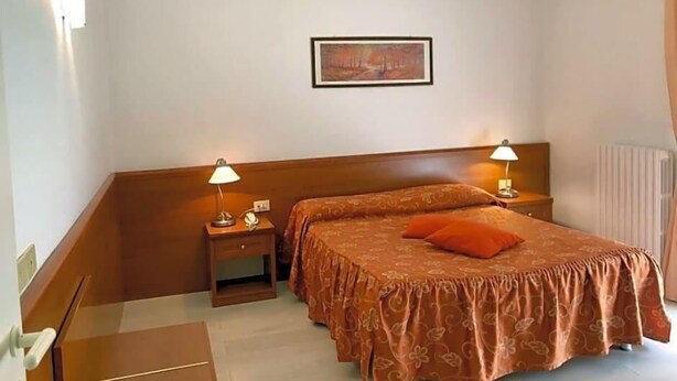 A hotel room with a bed, two bedside tables, a nightstand, a lamp, a framed picture on the wall, and a radiator. - Hotel Arcadia (Macerata)