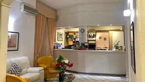 A hotel lobby with a reception desk, a seating area with a yellow armchair, a white sofa, and a coffee table with flowers. - Hotel Arcadia (Macerata)