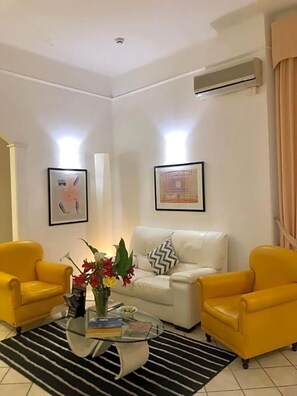 A living room with a white sofa, two yellow armchairs, a glass coffee table with books and flowers, a black and white striped rug, and two framed pictures on the wall. - Hotel Arcadia (Macerata)