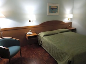 Habitación de hotel con cabecera de madera, colcha verde, una silla, mesita de noche con lámpara y un cuadro en la pared.
