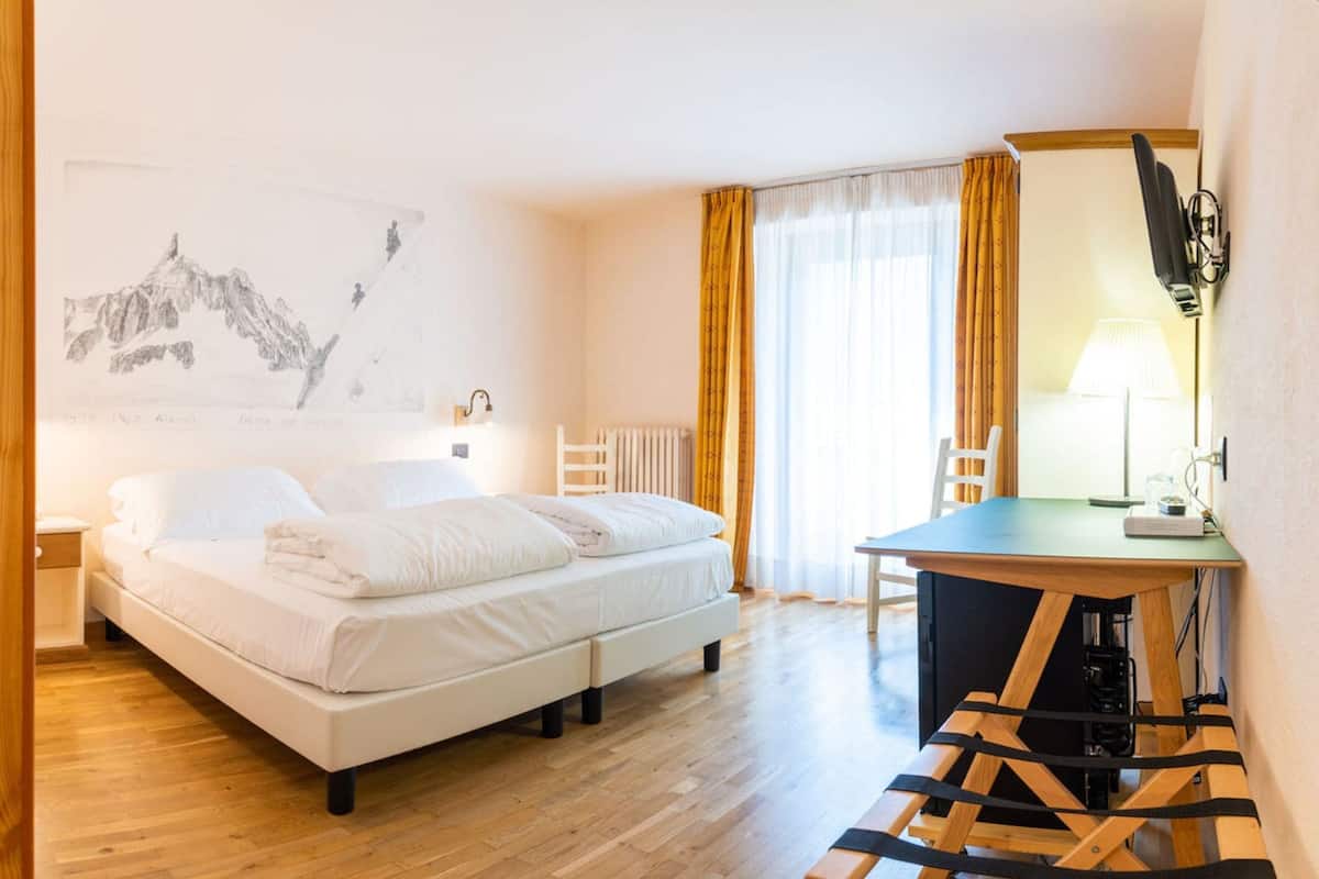 A hotel room with a bed, a desk, a chair, a radiator, and a wall-mounted TV.