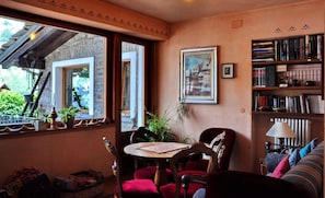 A cozy room with a wooden table, red chairs, a bookshelf, and a window overlooking a stone structure. - Berthod (Courmayeur)
