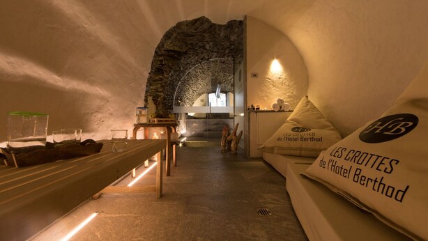 A dimly lit room with a stone archway, a bench, and a sofa with pillows displaying the hotel name.