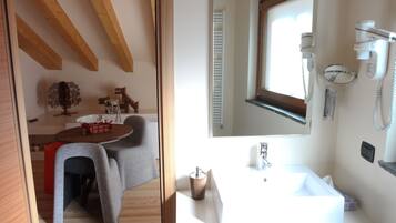 Baño con lavamanos blanco, espejo y secador de pelo. Hay una ventana sobre el lavamanos y una puerta que da a un comedor.