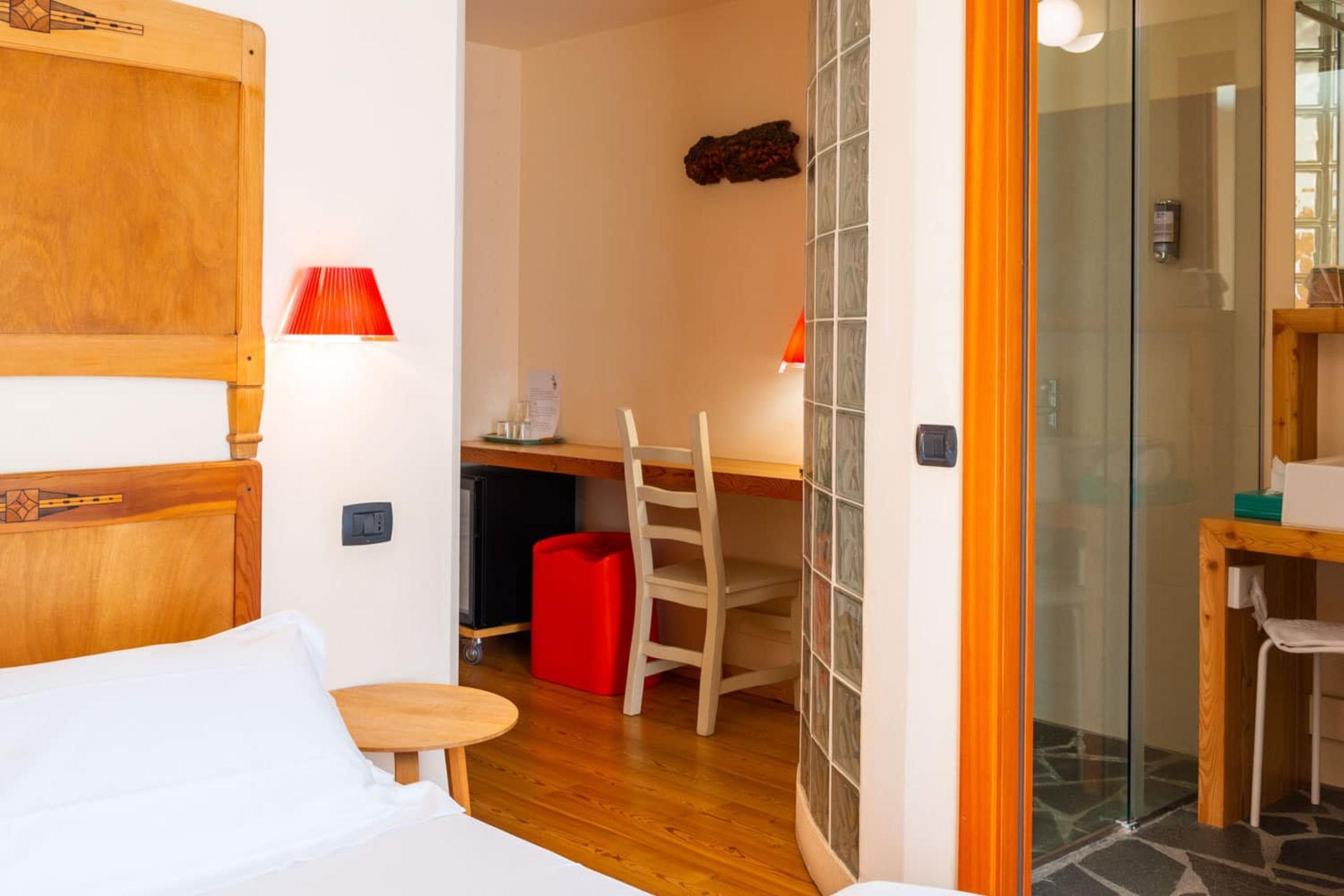 Quarto de hotel com cama, escrivaninha, cadeira, mesinha pequena, um cesto de armazenamento vermelho e box de vidro.