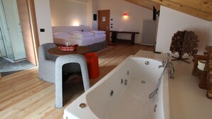 A modern, well-lit room with a wooden ceiling, a large bathtub, a cozy seating area, and a bed with white linens.
