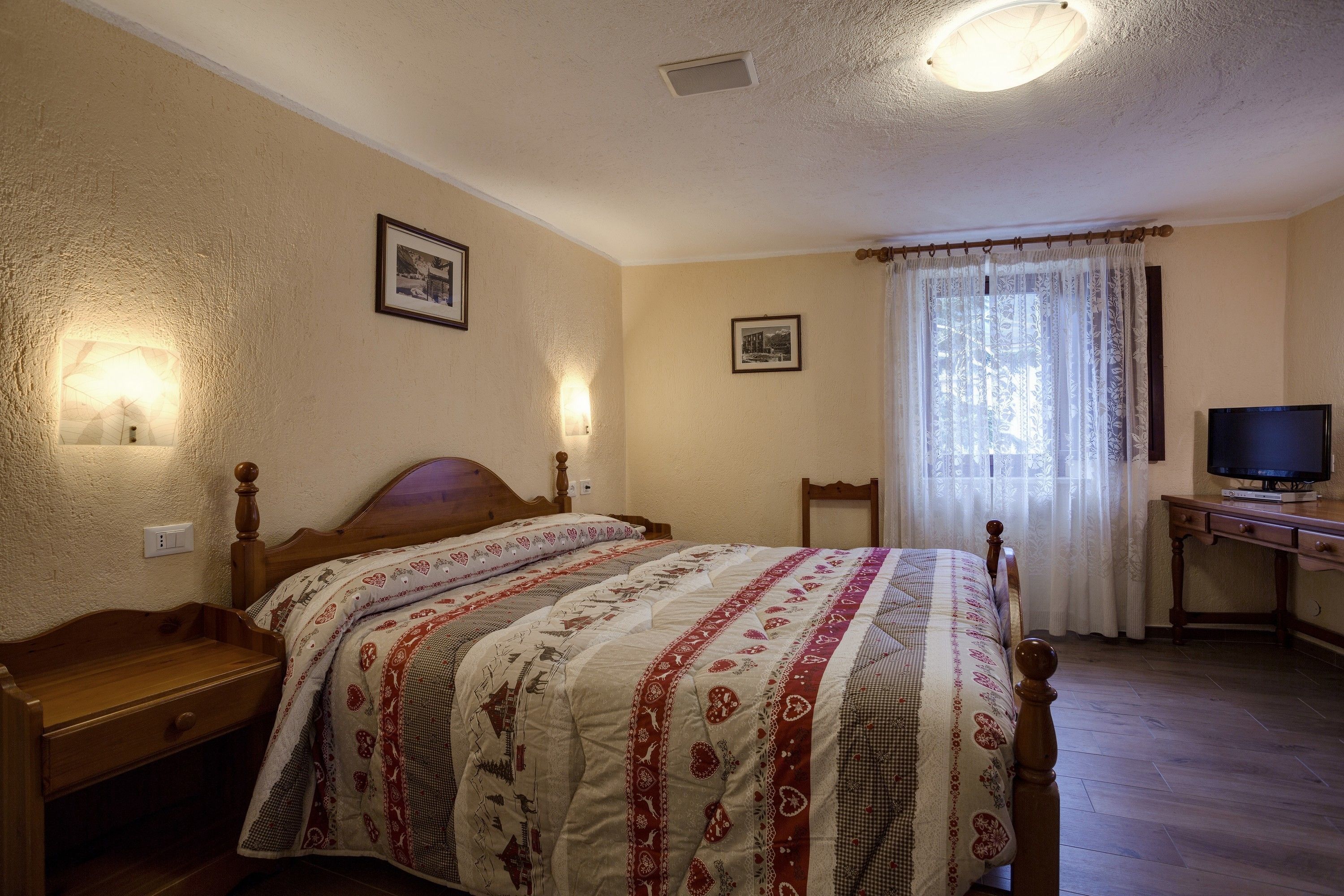A bedroom with a wooden bed, a nightstand, a desk with a television, and two wall-mounted lamps.