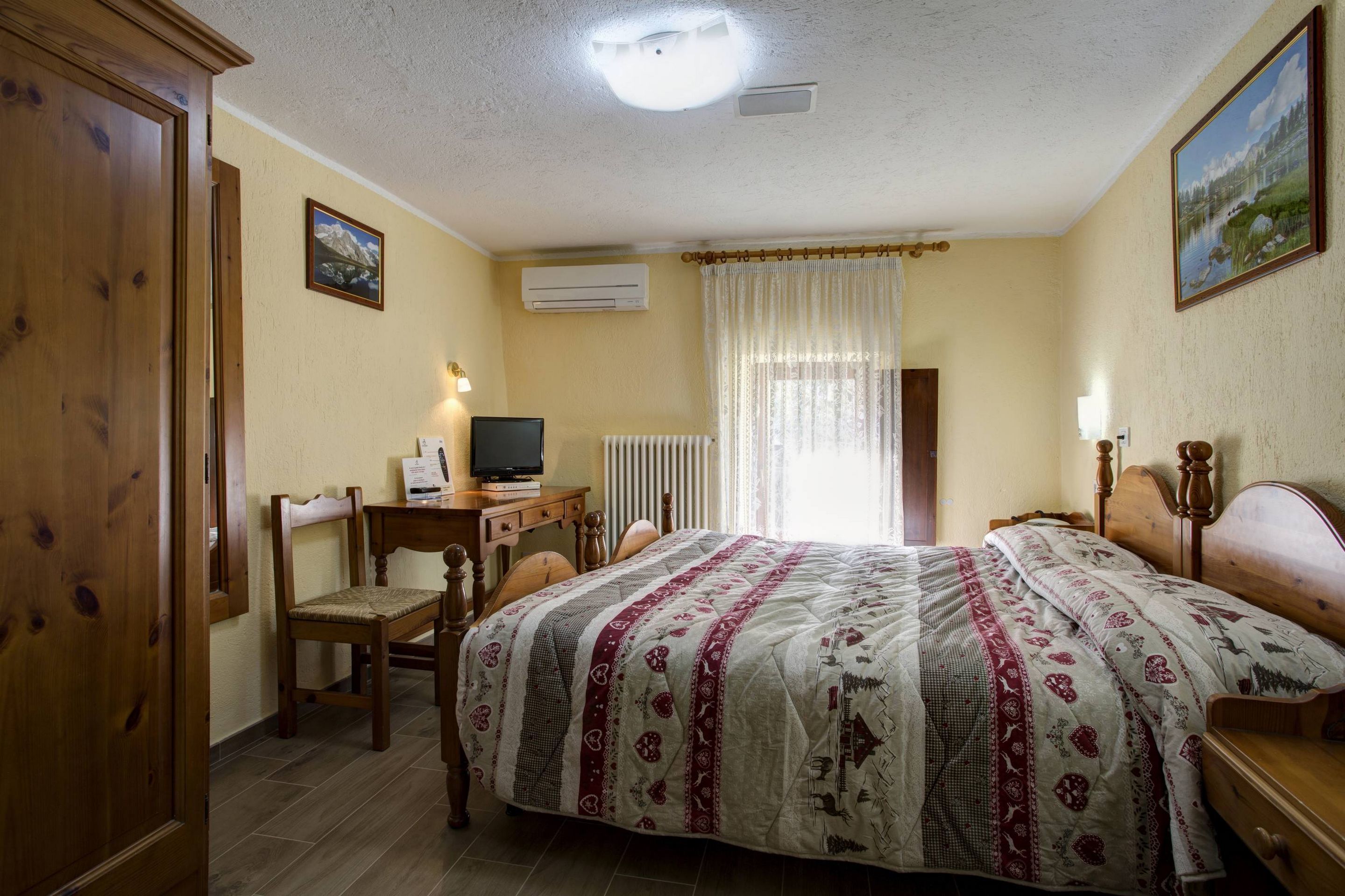 A hotel room with a bed, a desk with a computer, a chair, a radiator, and a window with curtains.