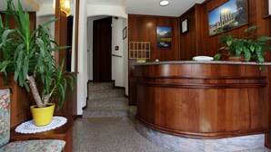 A reception area with a wooden reception desk, a potted plant, and a framed picture on the wall. - Hotel Cecchin (Aosta)