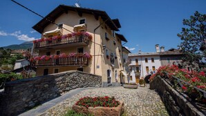 A traditional stone-paved street with a building featuring a balcony with red flowers, stone walls, and a clear blue sky. - Hotel Cecchin (Aosta)