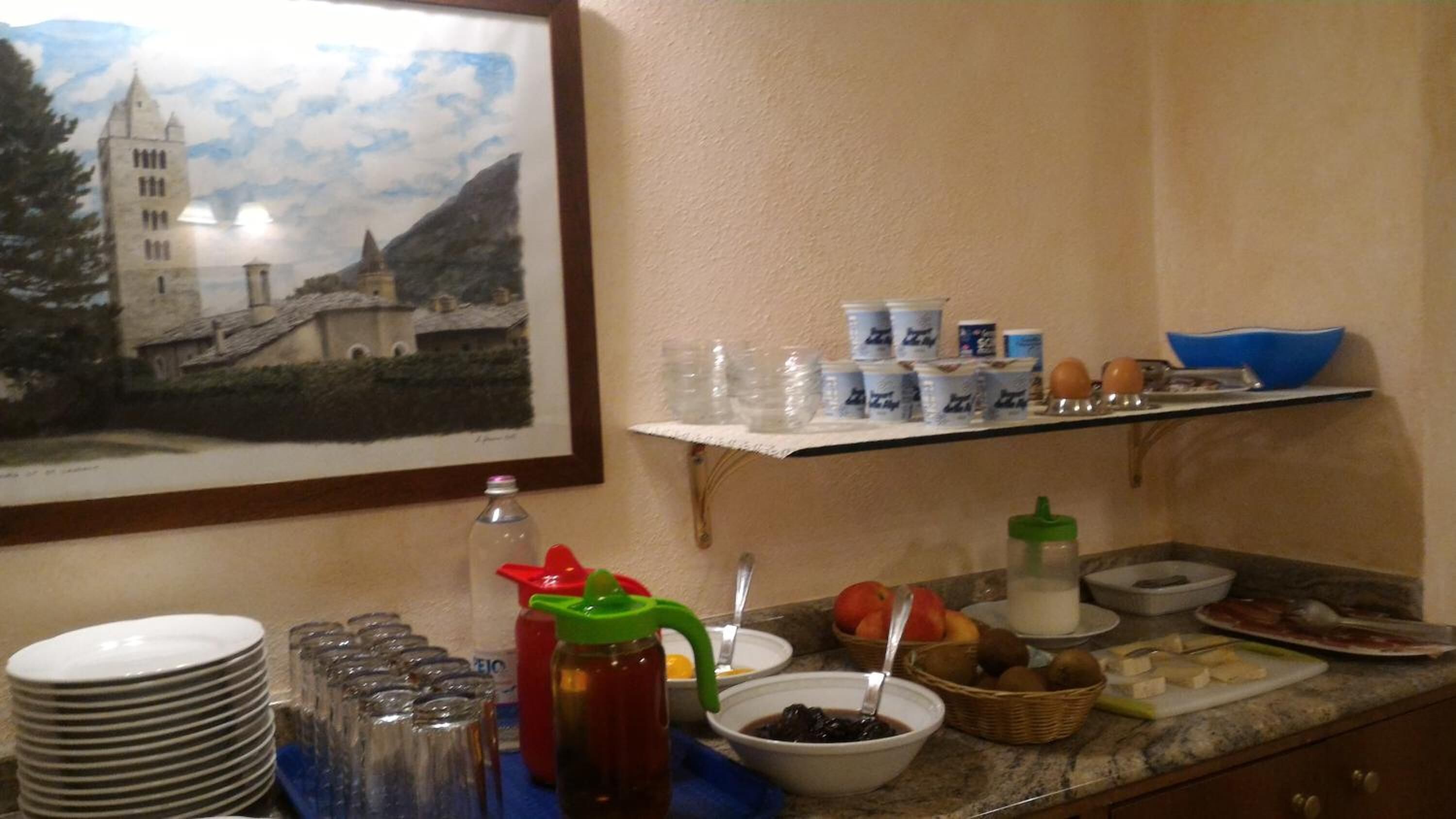 Un bufé de desayuno con diversos alimentos y bebidas, un cuadro enmarco de un edificio y una estantería con tazas y cuencos.