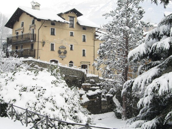 A snow-covered hotel with a balcony and a stone wall in the foreground. - Hotel Cecchin (Aosta)