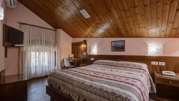 Um quarto com teto de madeira, cama com colcha estampada, escrivaninha com cadeira, televisão e janela com cortinas.