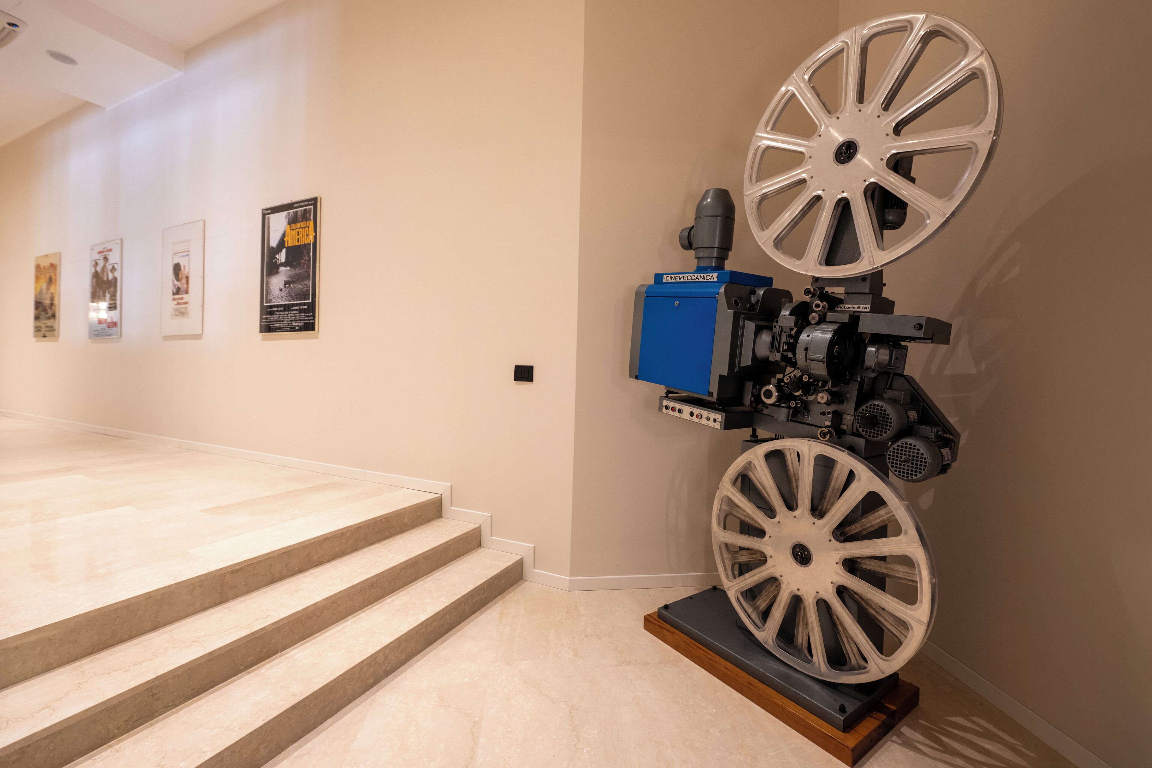 A modern interior with a staircase leading to a higher floor, a wall-mounted projector, and framed movie posters.