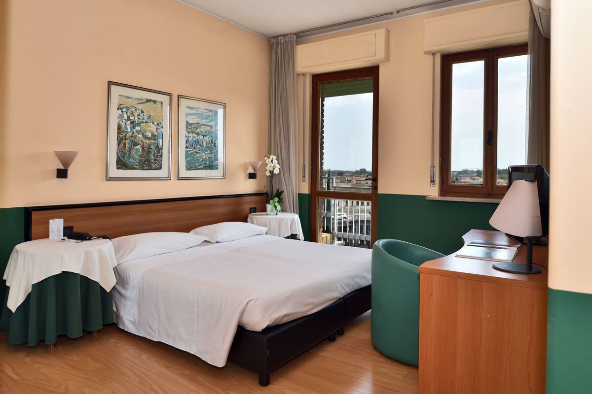 A hotel room with a bed, a desk, a chair, a window with a view, and two paintings on the wall.