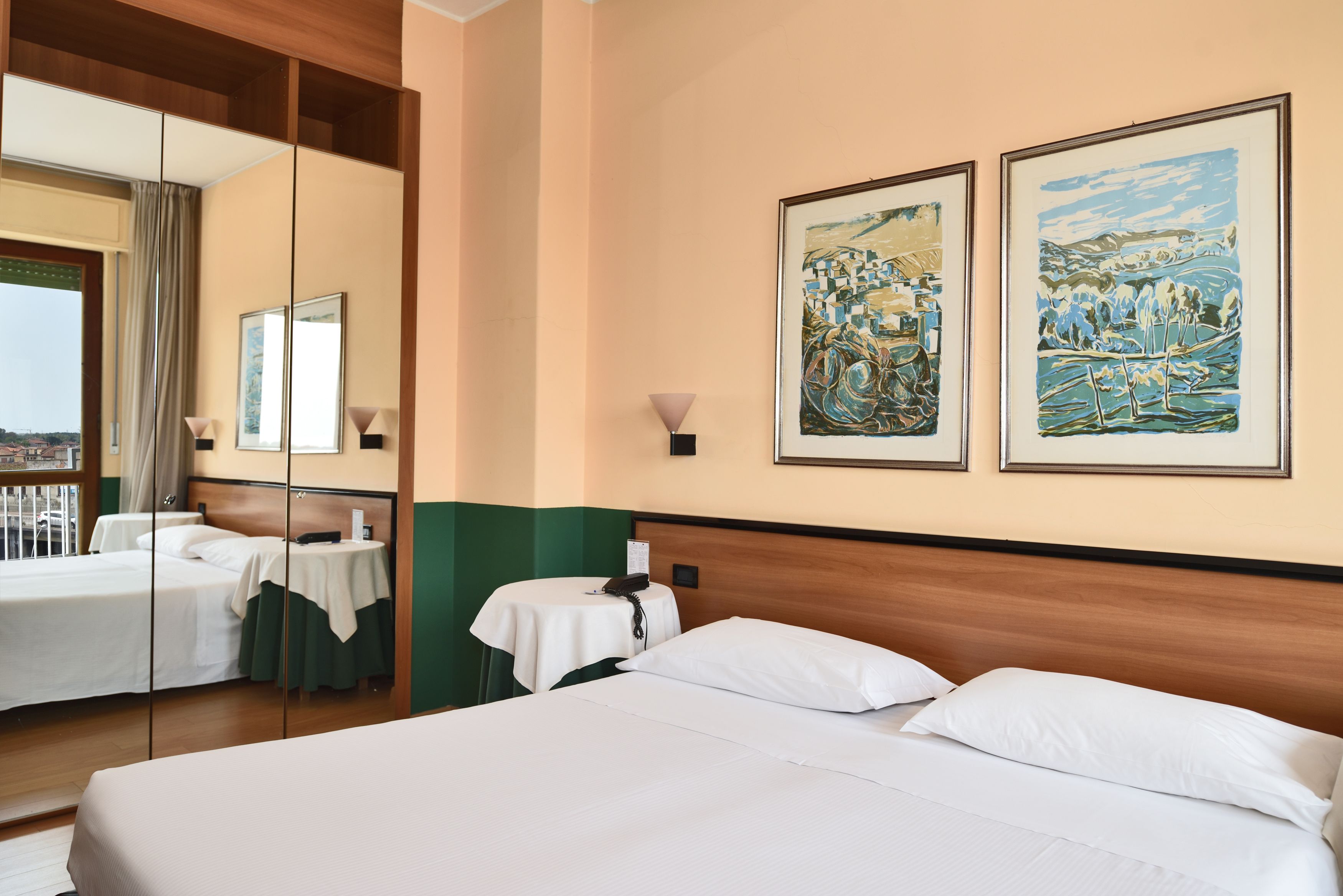 Una habitación de hotel con una cama grande, una mesita de noche con lámpara, una pequeña mesa con teléfono y dos obras de arte enmarcadas en la pared.