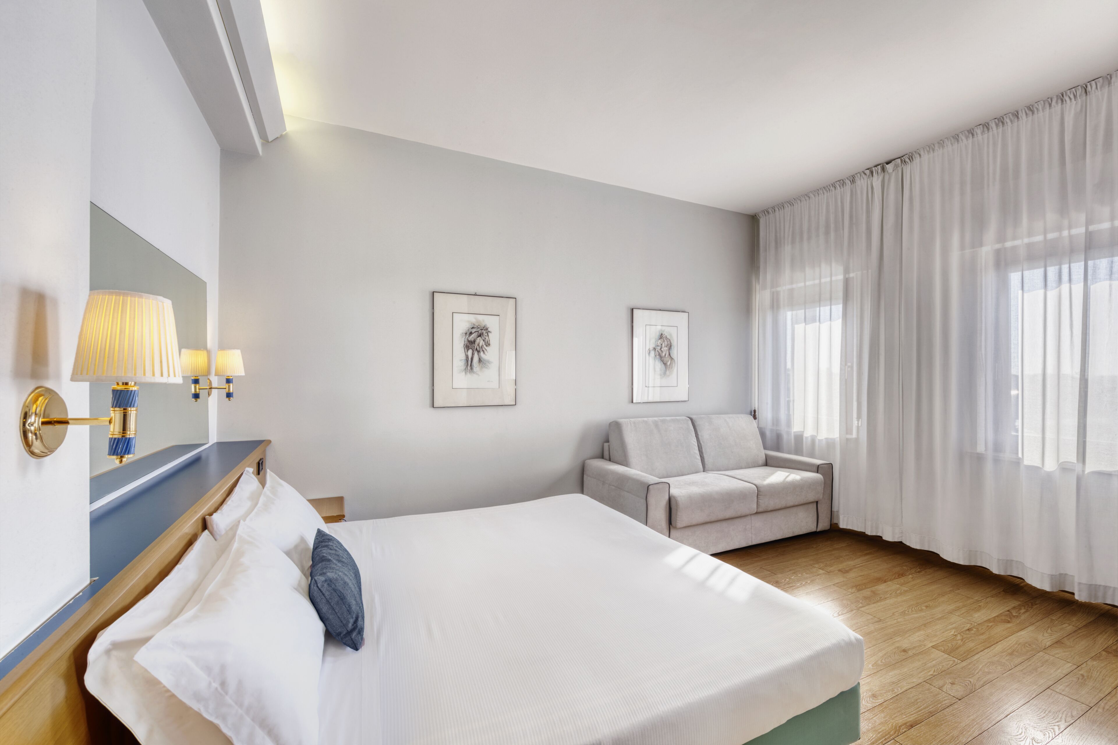 Una habitación de hotel con una cama, un sofá, una lámpara fijada a la pared y dos cuadros enmarcados.