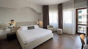 A modern hotel room with a large bed, a sitting area with a chair, a small table, and a balcony with a view.