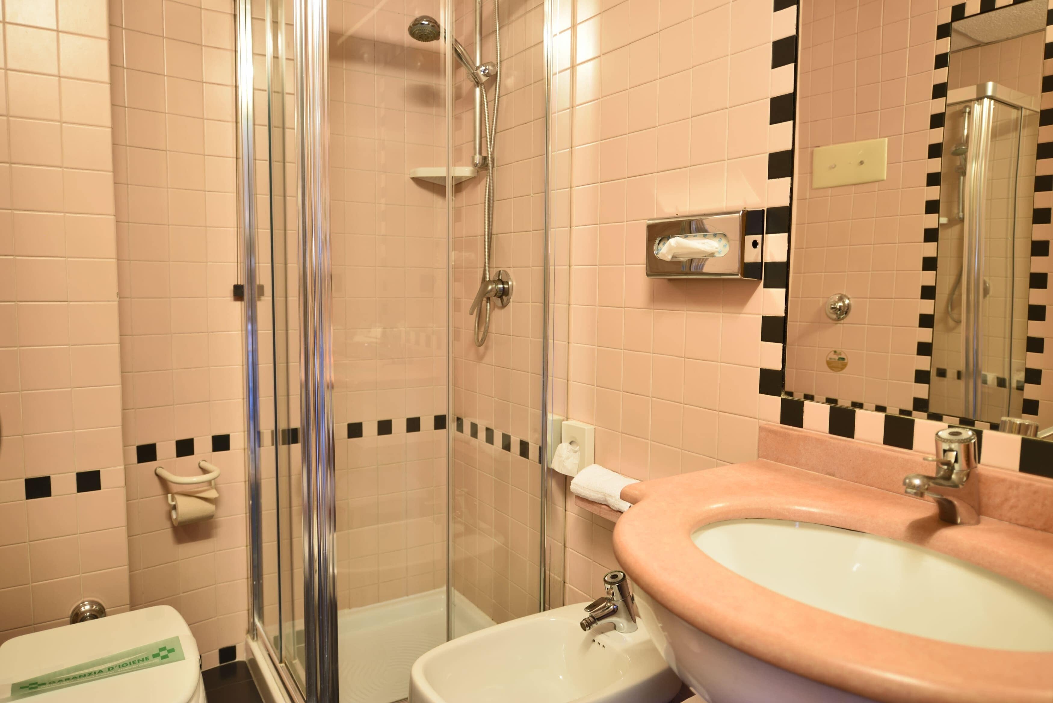 A bathroom with a glass-enclosed shower, a sink with a pink basin, and a toilet with a green sign.