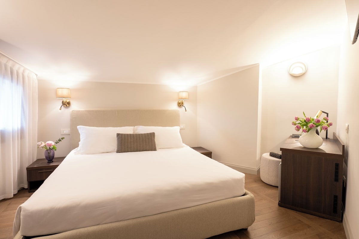 A neatly made bed with a headboard, two bedside tables with lamps, a small round ottoman, and a cabinet with a vase of flowers.