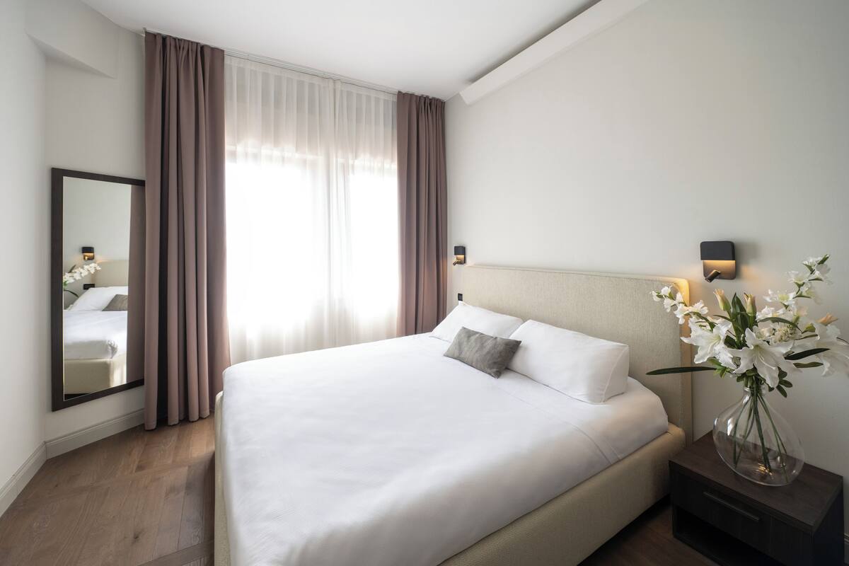 A neatly made bed with white linens, a nightstand with a vase of flowers, and a large window with sheer curtains.