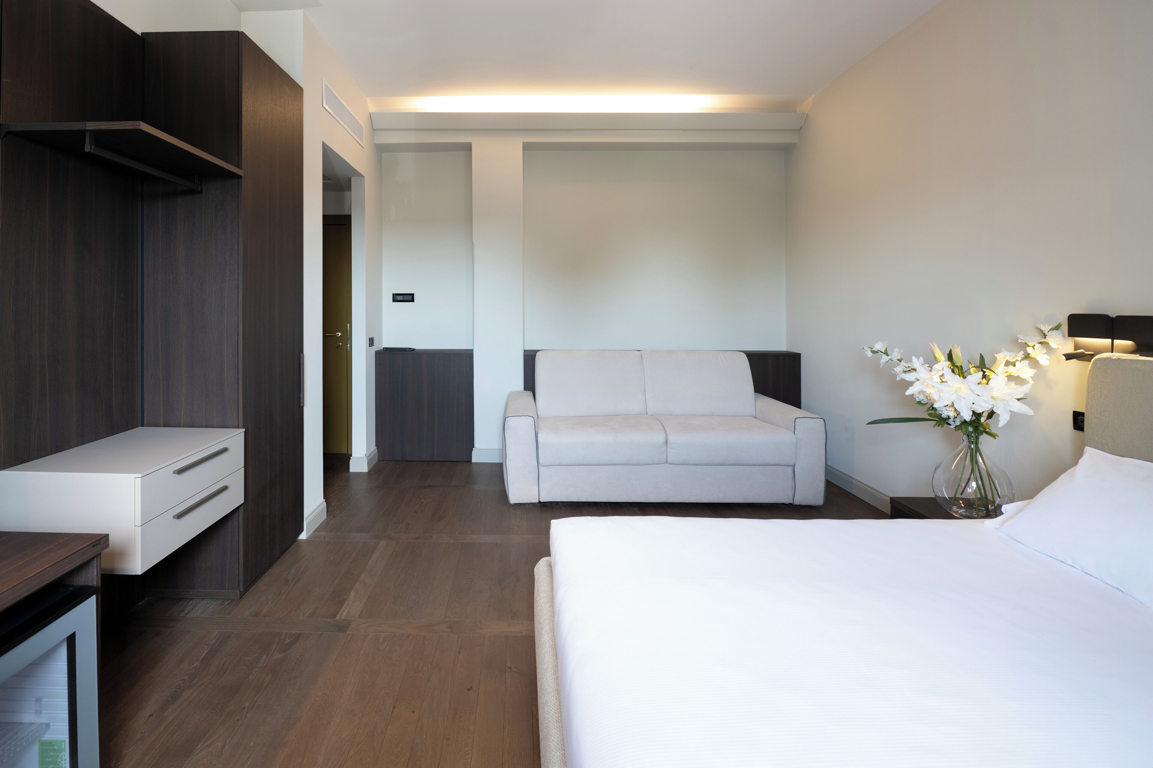 A modern hotel room with a bed, a sofa, a desk, and a nightstand.