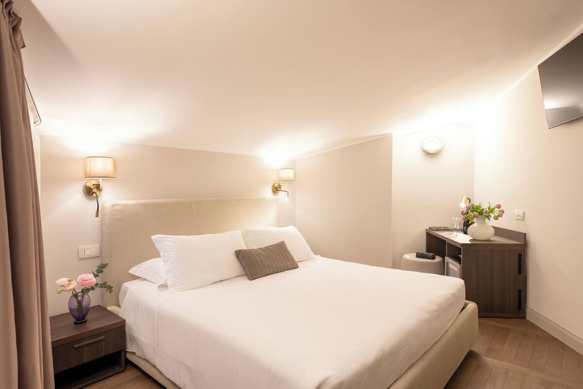 A neatly made bed with white linens and two pillows, a bedside table with a vase of flowers, and a small desk with a lamp.