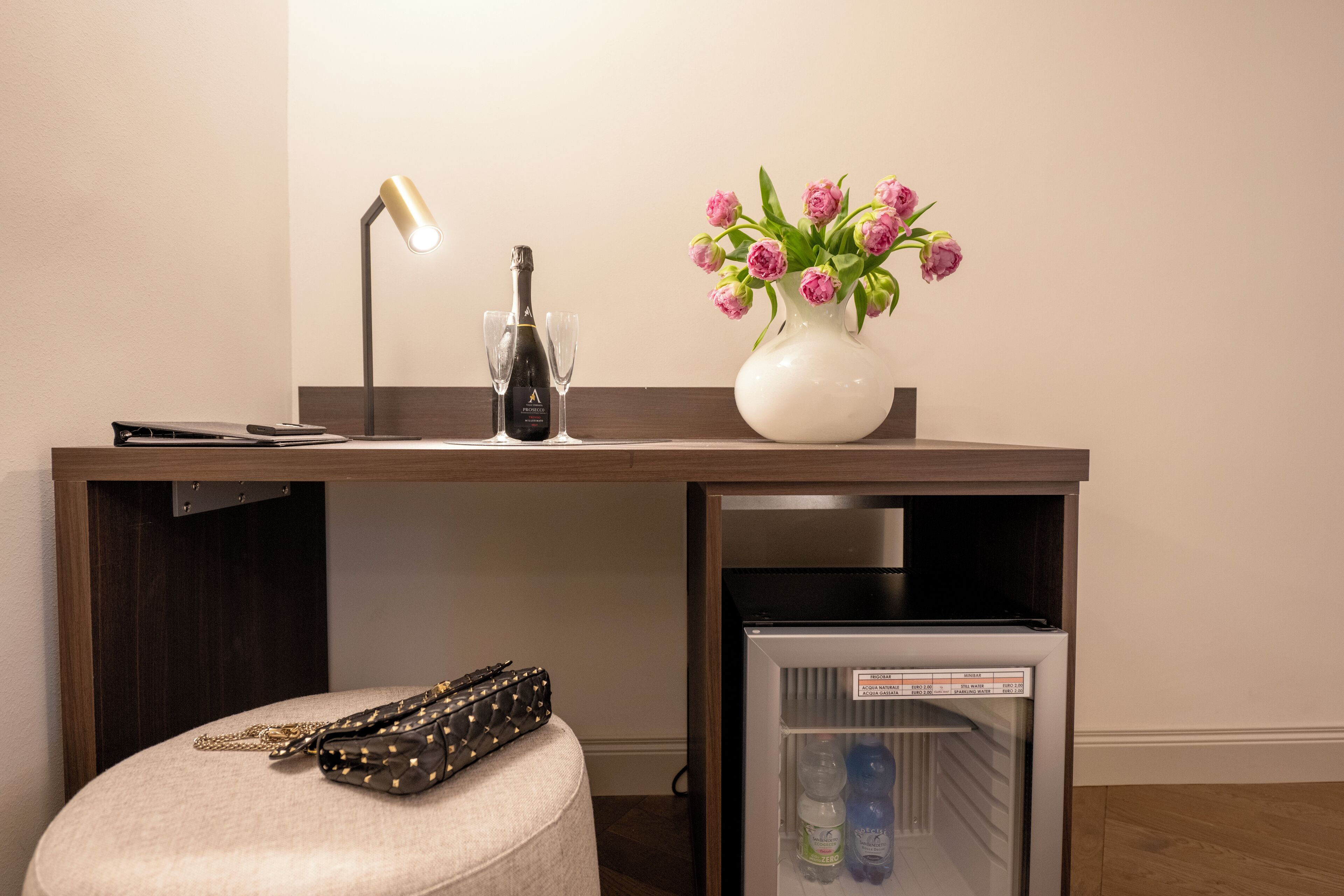 Un bureau compact avec une lampe, une bouteille de champagne, deux verres, un vase avec des fleurs roses et un petit réfrigérateur.
