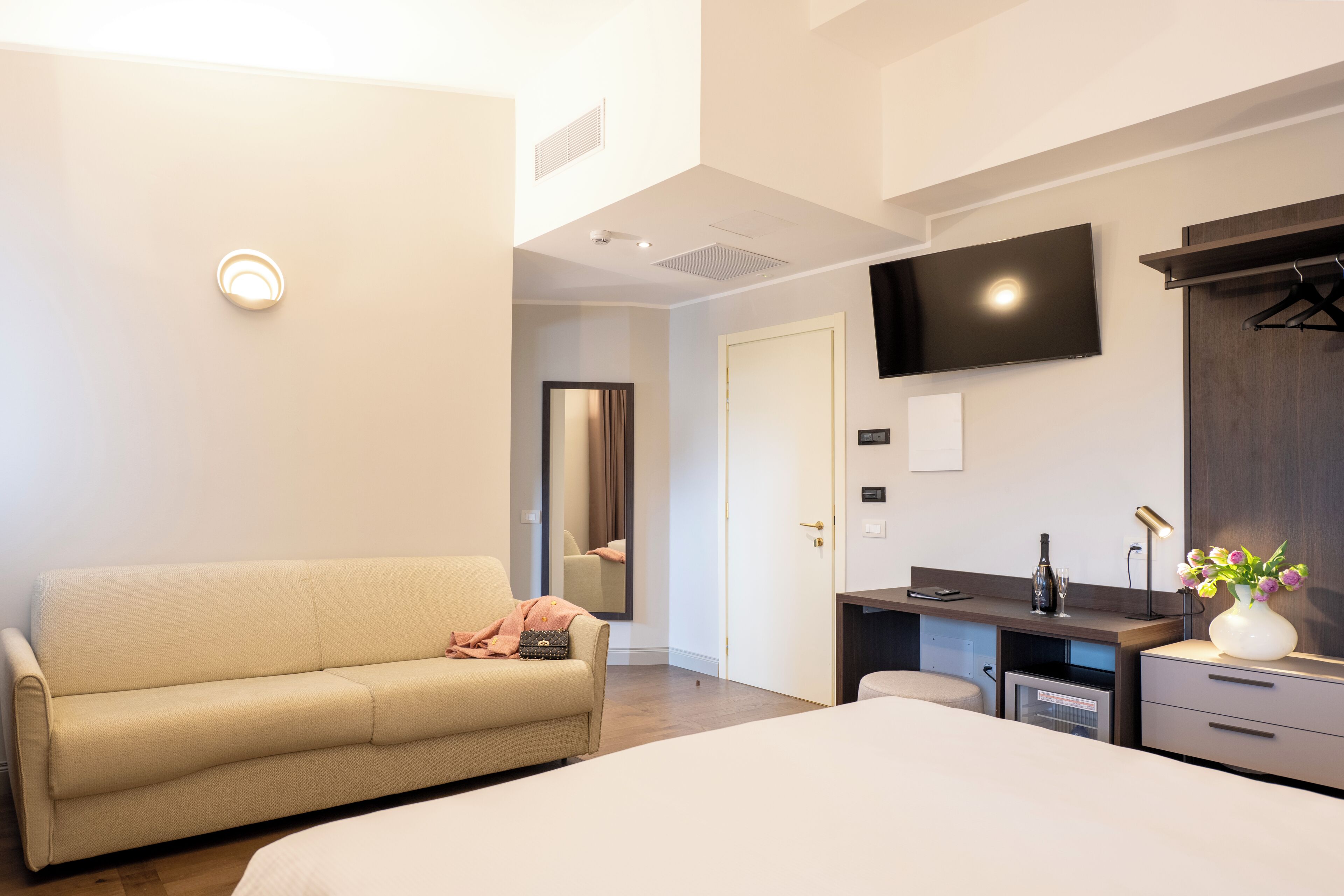 A modern hotel room with a bed, a sofa, a desk with a chair, a flat-screen TV, and a mirror.