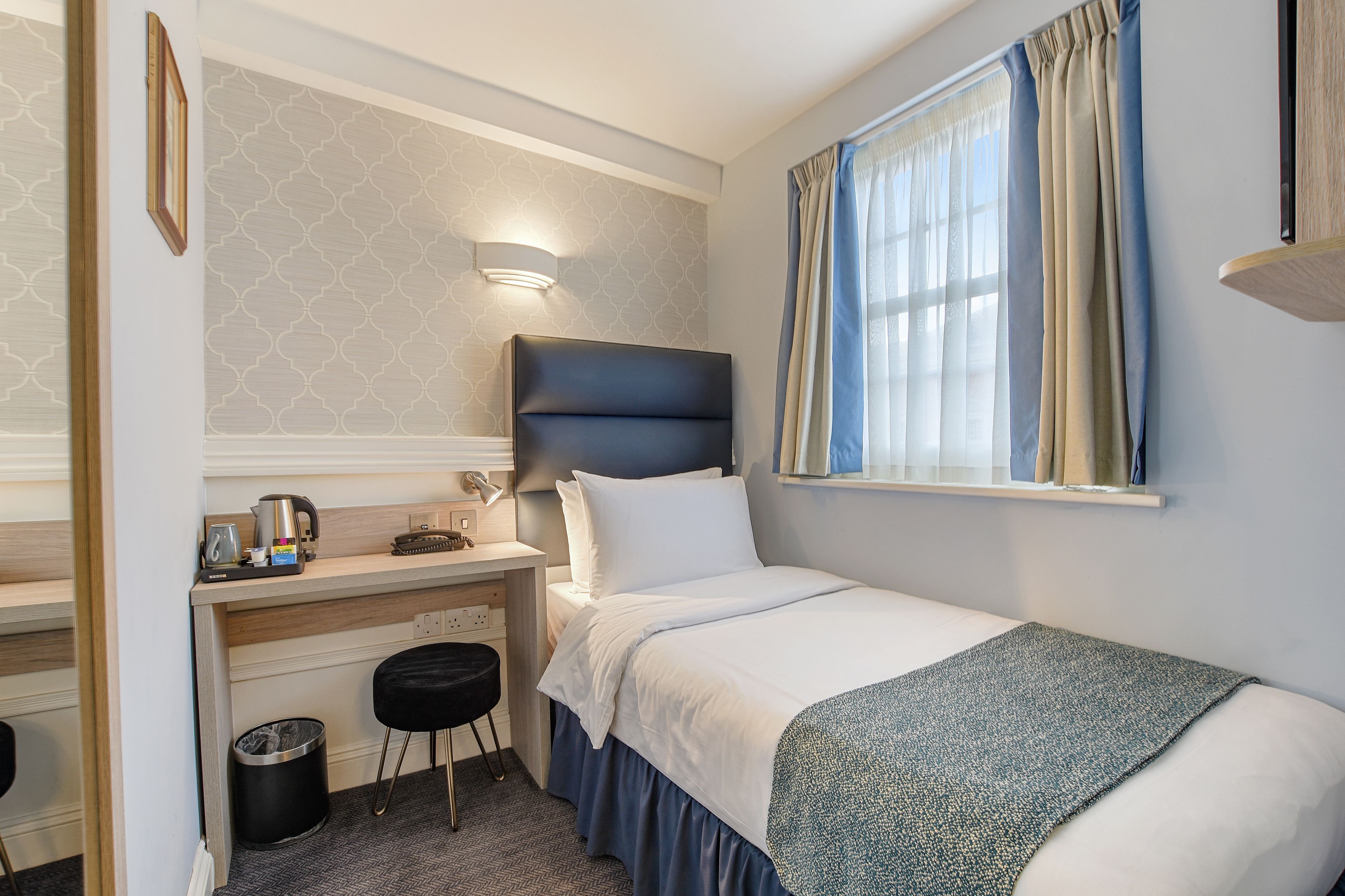 A compact hotel room with a single bed, a desk with a chair, a small trash bin, and a wall-mounted light fixture.