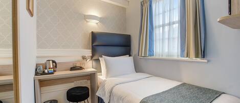A compact hotel room with a single bed, a desk with a chair, a small trash bin, and a wall-mounted light fixture.