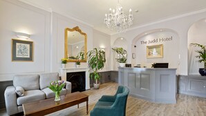A hotel lobby with a grey sofa, teal armchairs, a wooden coffee table, a chandelier, a fireplace, and a reception desk.