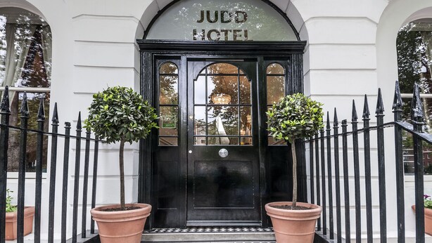 A black entrance door with the sign "JUDO HOTEL" above it, flanked by two potted plants. - Judd Hotel (London)